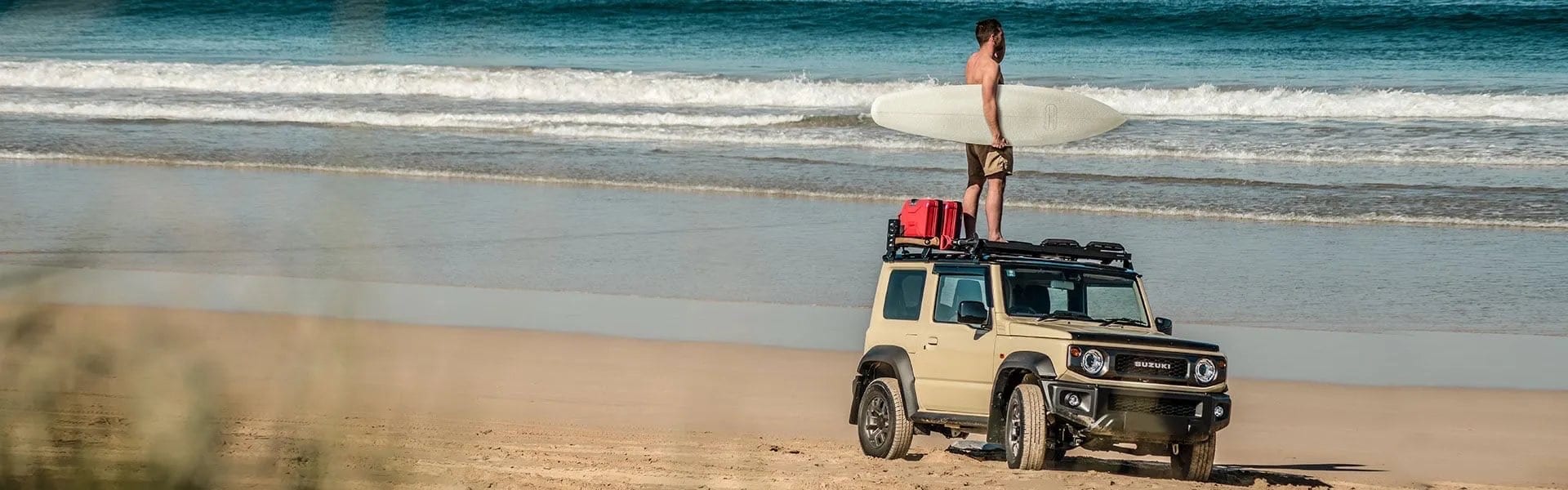 Beige Suzuki Jimny 3-door on a beach with a surfer standing on its roof rack