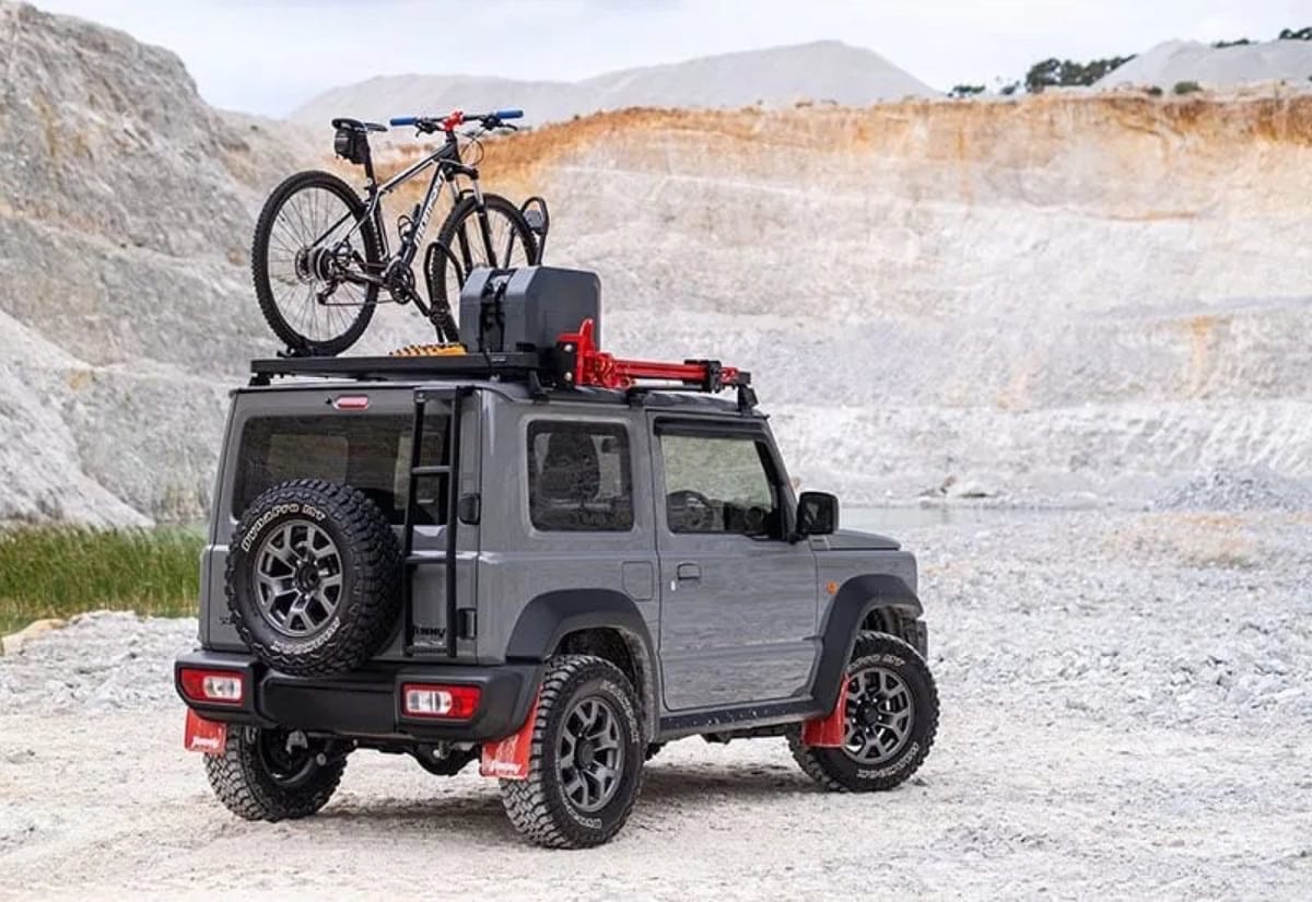 Grey Jimny 3-door with a roof rack, bike carrier and rear ladder in a quarry