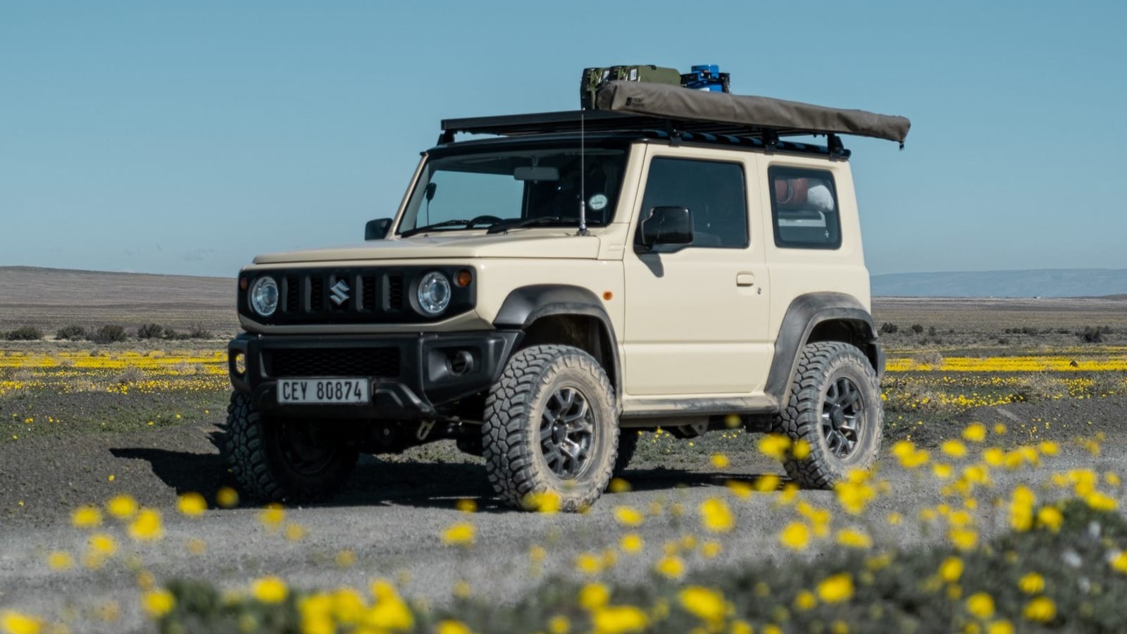 A beige current Suzuki Jimny 3-door with a roof rack and chunky all-terrain tyres, parked on West Coast gravel behind yellow spring flowers