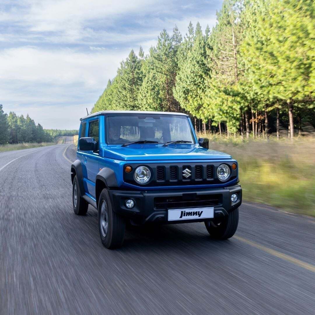 A blue current Suzuki Jimny 3-door driving on a tar road through pine plantations