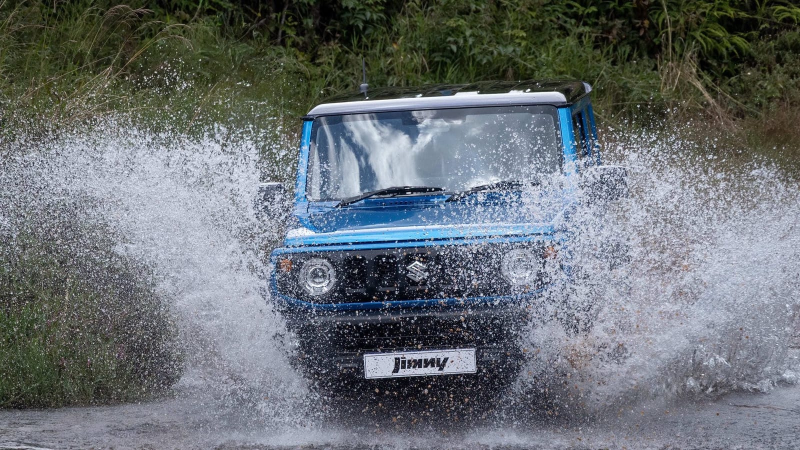 A blue standard-looking Suzuki Jimny 3-door driving head-on through a shallow water crossing with a big splash, bush behind