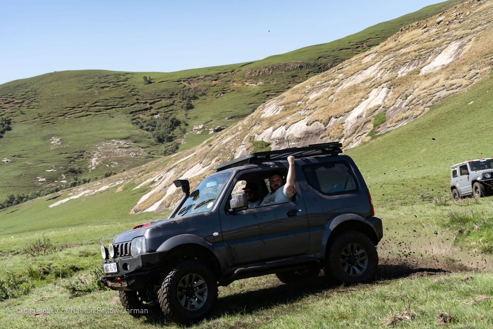 A dark grey older Suzuki Jimny with a snorkel and mud-terrain tyres throwing turf off its rear wheel on a steep green slope