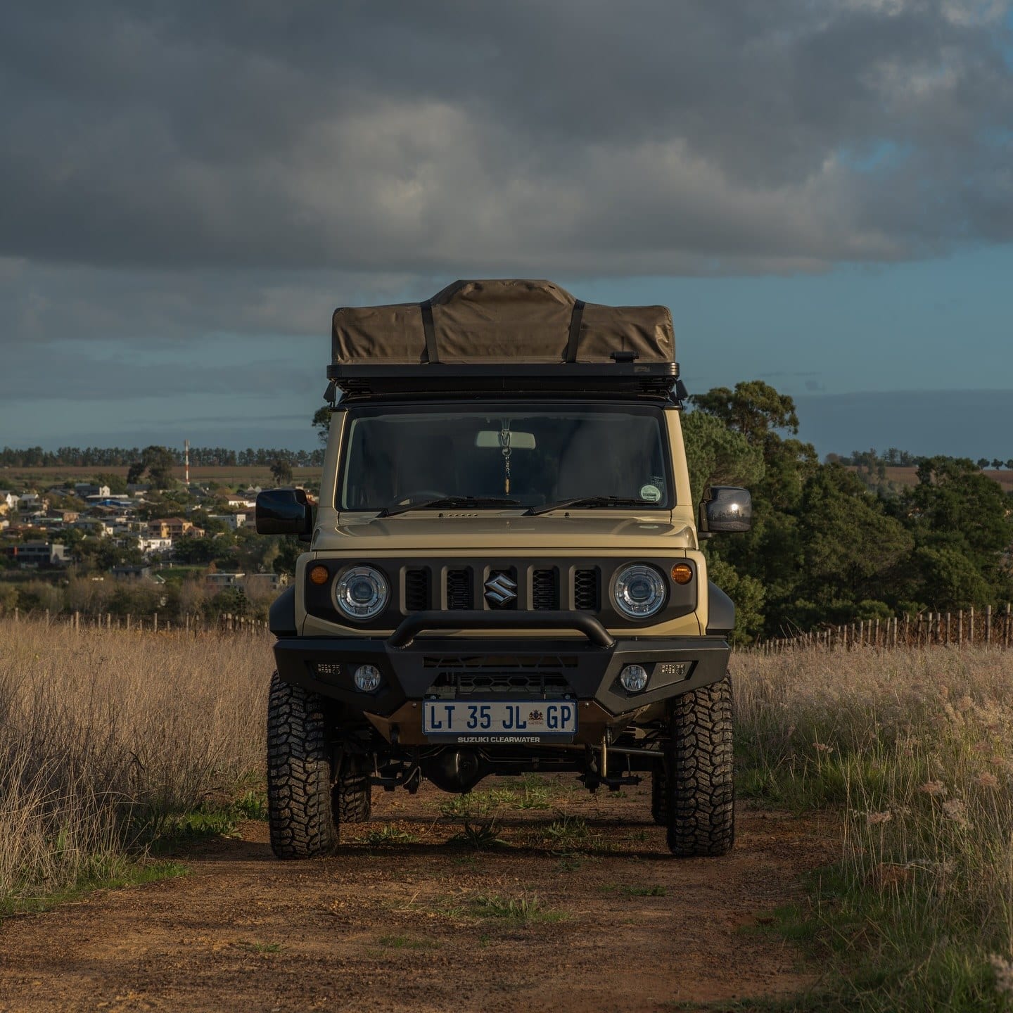A sand-coloured current Suzuki Jimny 3-door with a rooftop tent and chunky tyres, head-on on a dirt road with grassland behind