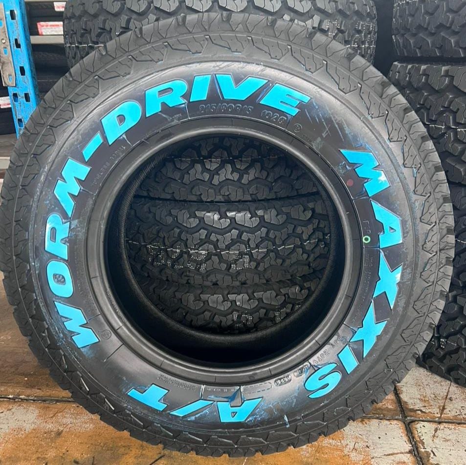 A Maxxis Worm Drive all-terrain tyre standing upright in a tyre shop, with bright blue sidewall letters and a stack of tyres behind it