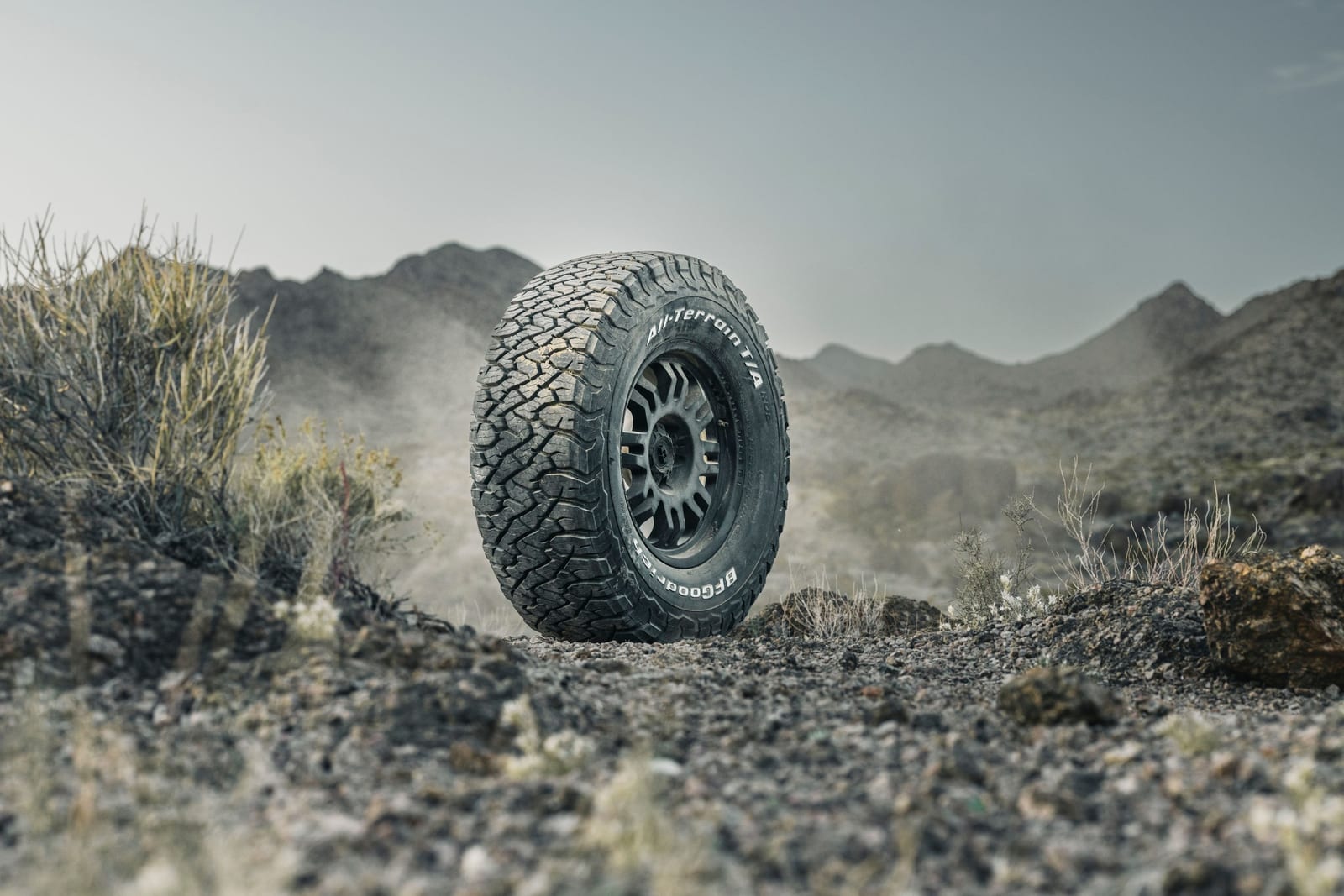 A single BFGoodrich KO3 all-terrain tyre on a dark wheel standing on stony ground, with dust and mountains behind