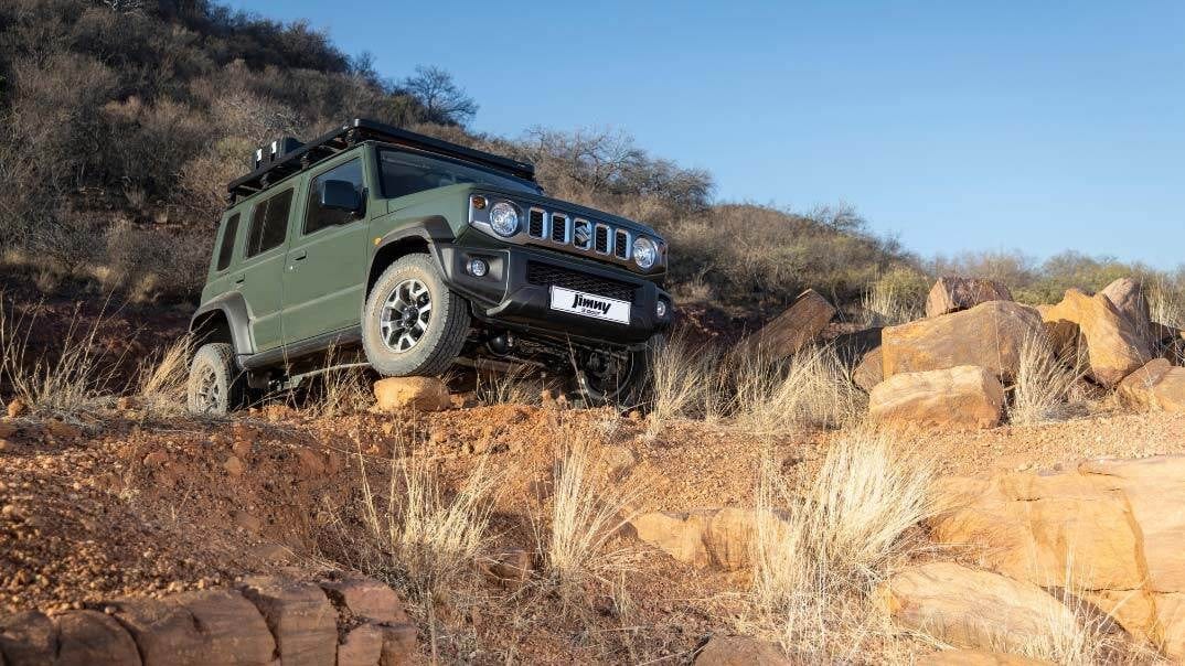 A green current Suzuki Jimny 5-door with a roof rack climbing over red rock and dry grass on its factory wheels and tyres