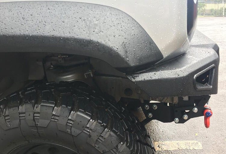 Close-up of the front corner of a Suzuki Jimny with a steel bumper whose short end sits clear of a big off-road tyre, with a red recovery shackle
