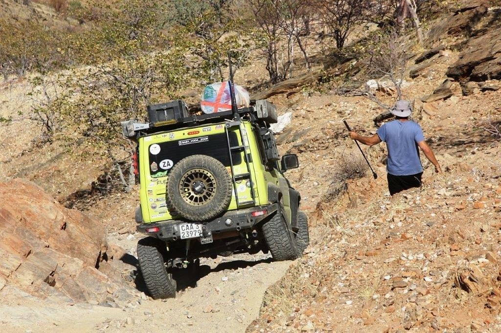 A lime green Suzuki Jimny seen from behind climbing a rocky gully, with one rear wheel pushed up into the arch while a man walks ahead to spot