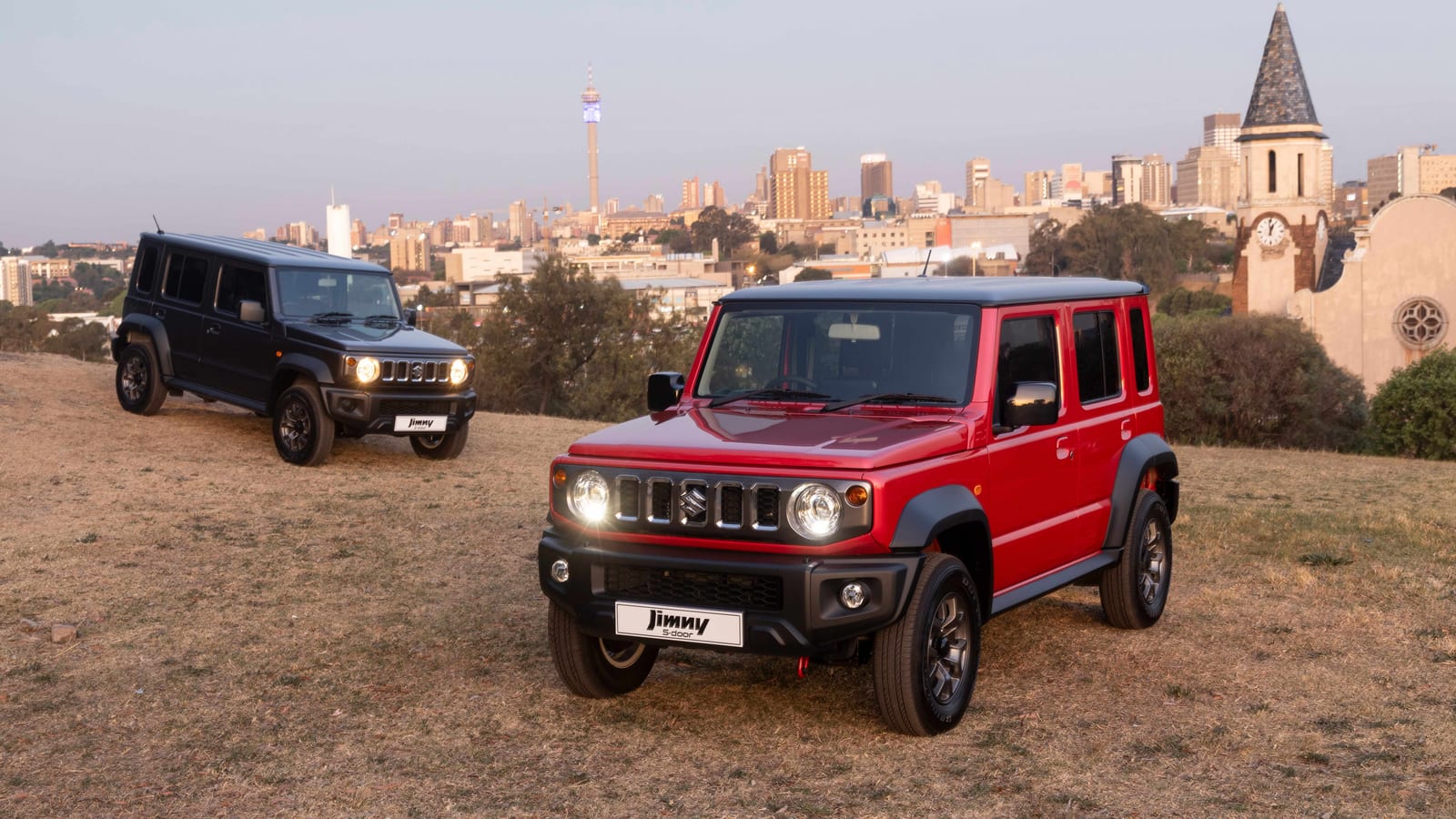 A red and a black Suzuki Jimny 5-door parked on a grassy hill with the Johannesburg skyline behind at dusk