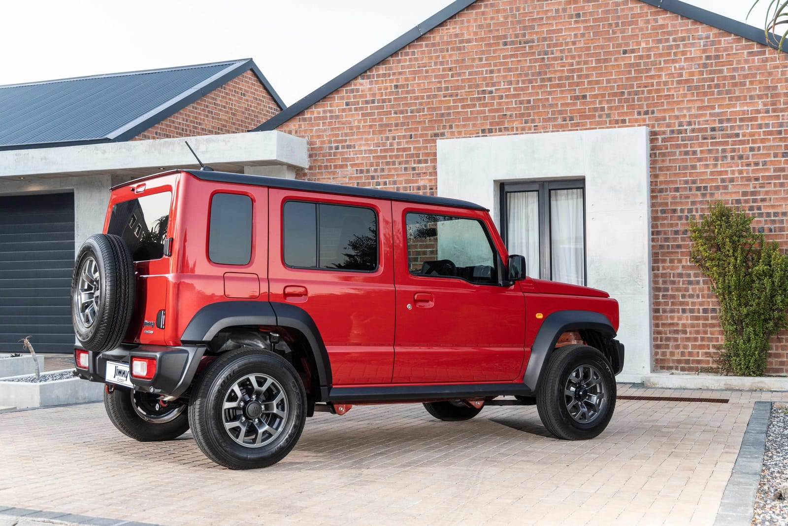 A red Suzuki Jimny 5-door parked in the paved driveway of a red-brick house