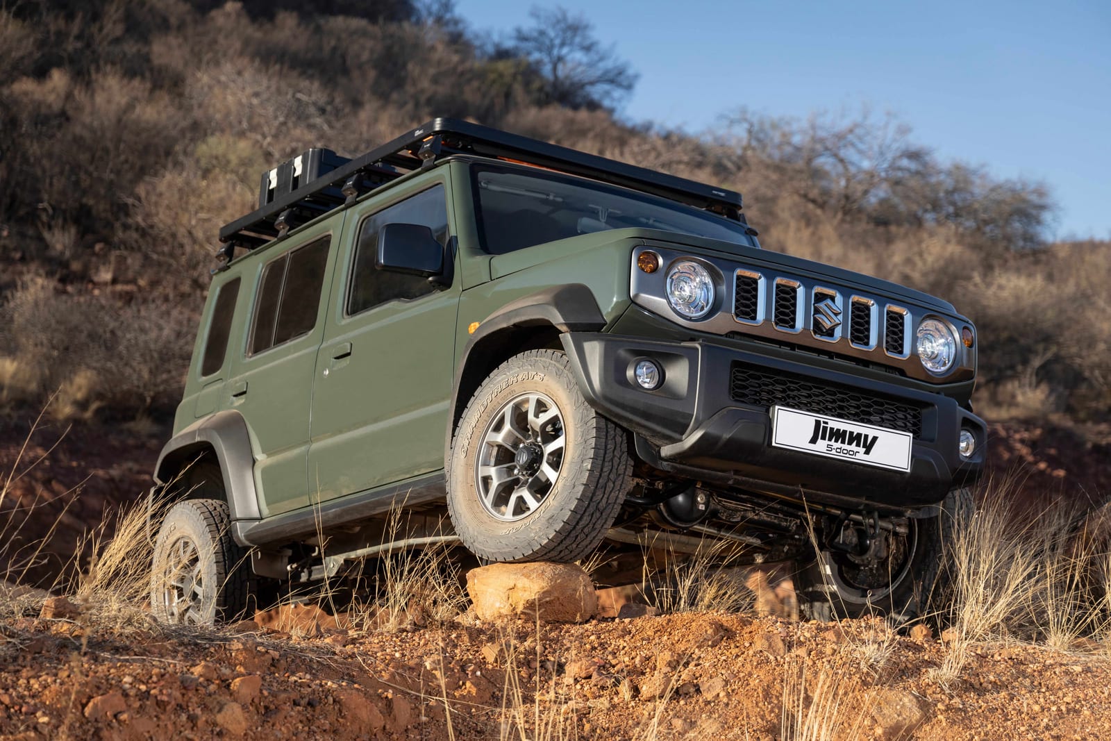 A green Suzuki Jimny 5-door with a roof rack and all-terrain tyres, one front wheel up on a rock in dry bush