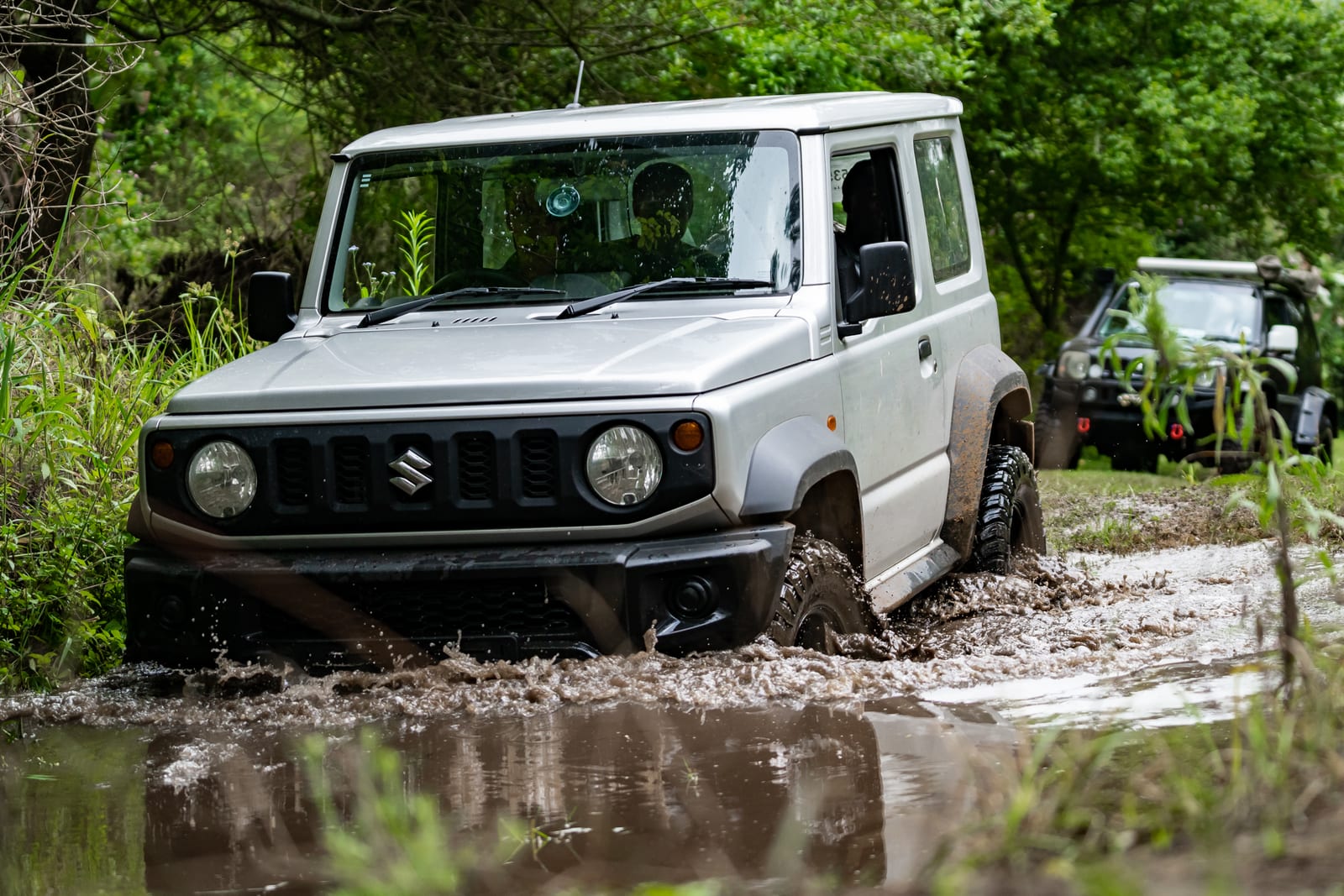 A lifted silver current Suzuki Jimny 3-door on mud tyres splashing through a muddy puddle, with an older Jimny parked behind it