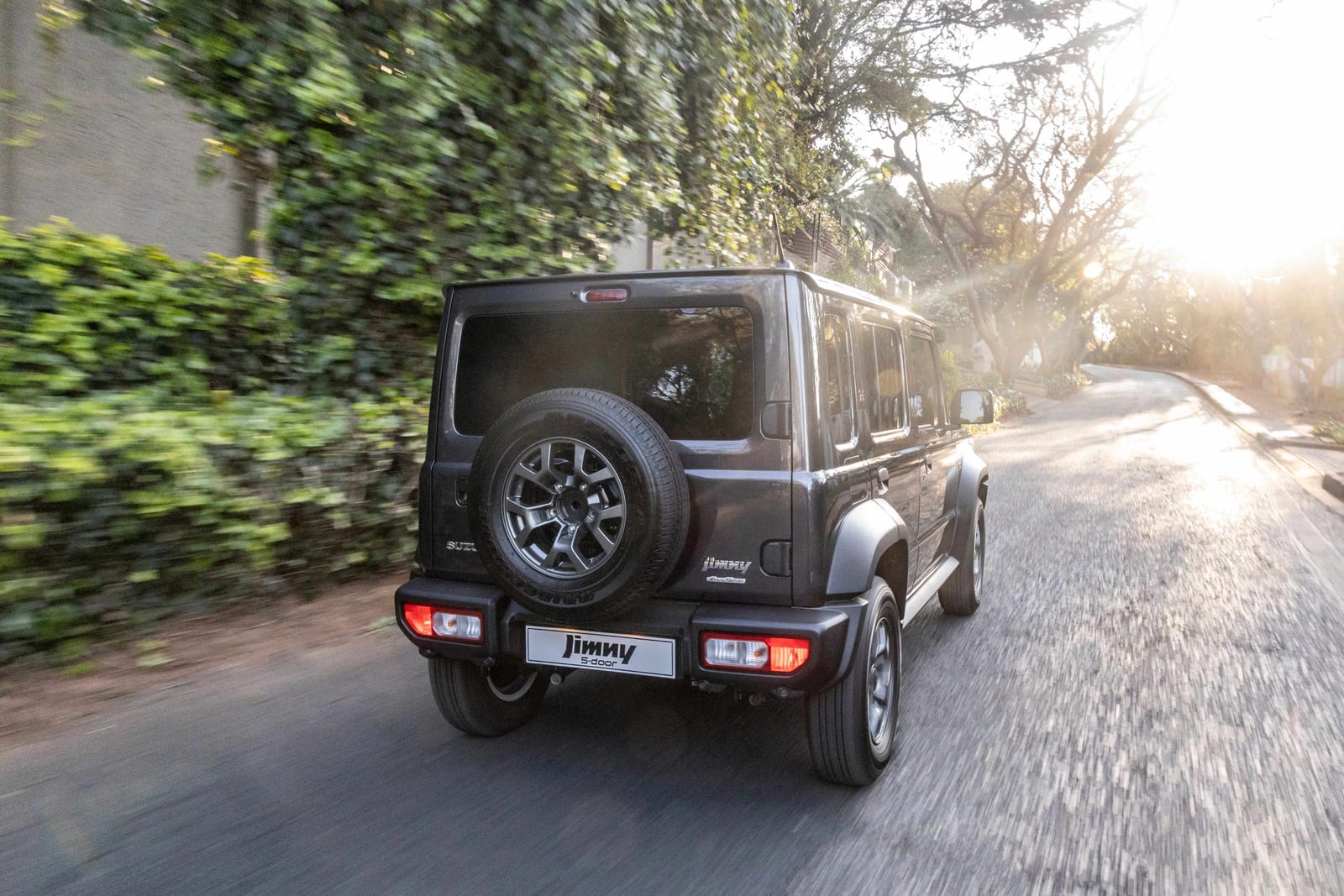 A dark grey Suzuki Jimny 5-door seen from behind, driving down a leafy suburban road in low sun
