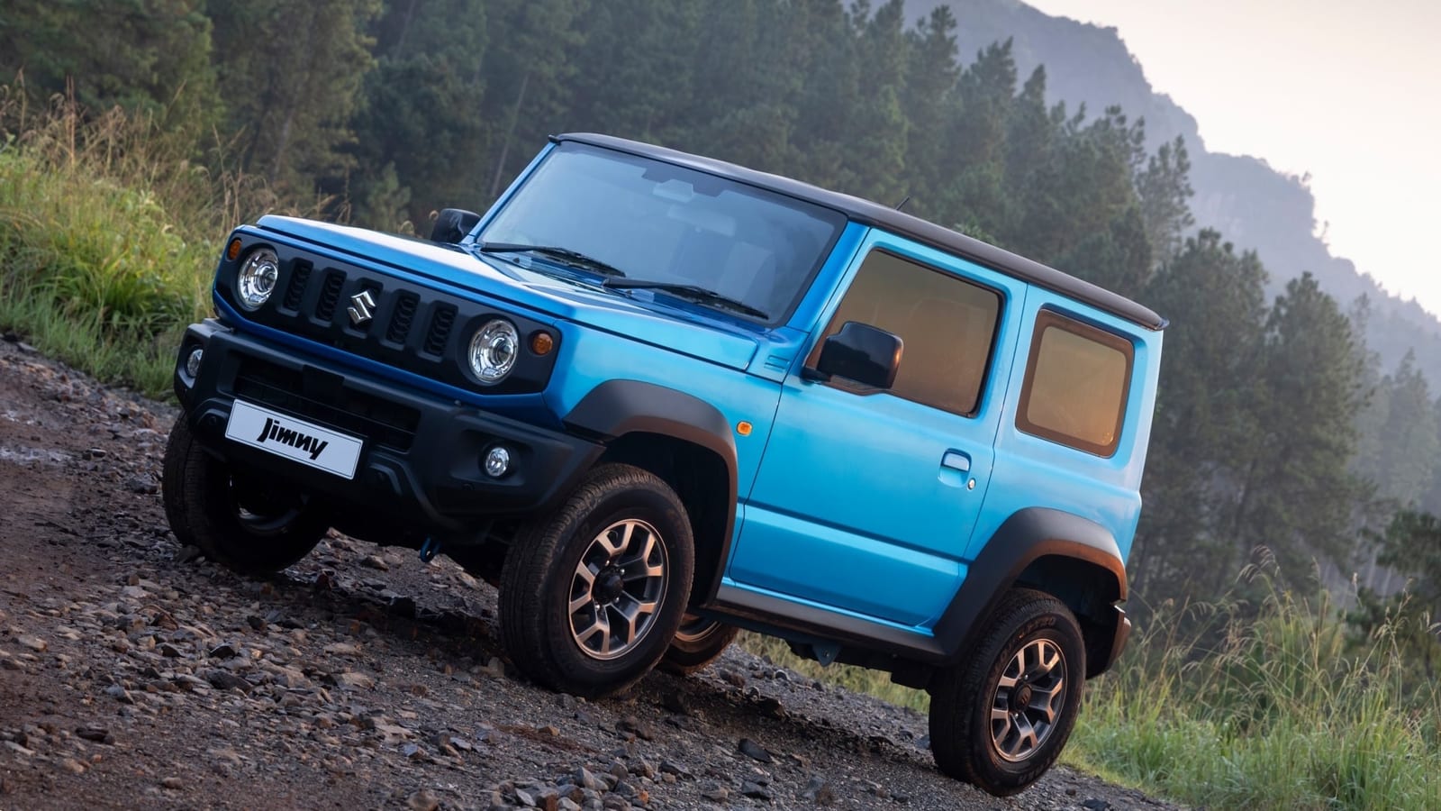 A standard blue Suzuki Jimny 3-door climbing a steep, stony track through pine forest