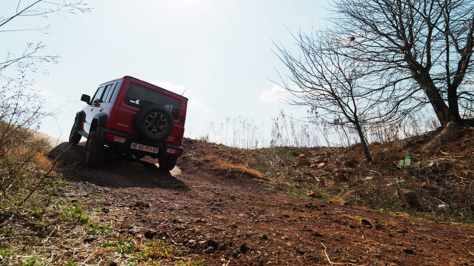 A standard red Suzuki Jimny 5-door from behind, climbing out over a dirt mound with the body leaning and the axles at different angles