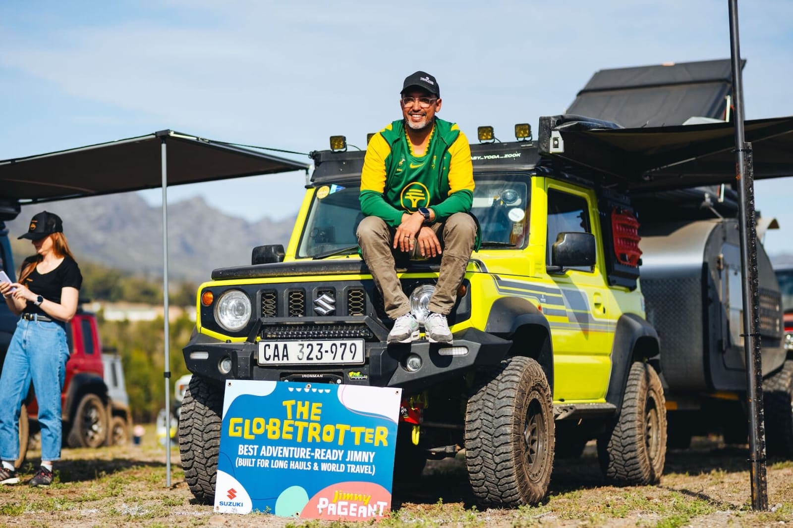 A young man in a green and gold jersey sits on the bonnet of a lime green Jimny 5-door at an outdoor gathering, with mountains behind