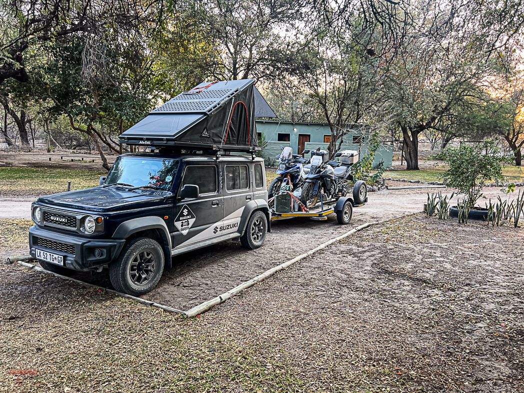 A Suzuki Jimny 5-door with a rooftop tent towing a trailer loaded with adventure motorbikes at a bushveld campsite
