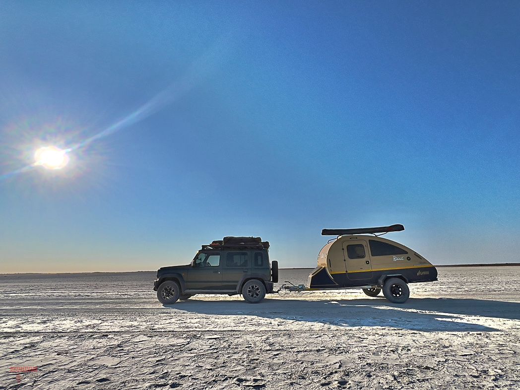 A Suzuki Jimny 5-door towing a teardrop camping trailer across a salt pan at sunset