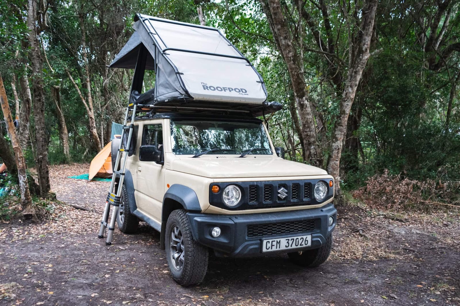 An ivory Suzuki Jimny 3-door parked in a forest camp with a RoofPod clamshell tent open on its roof and a ladder down
