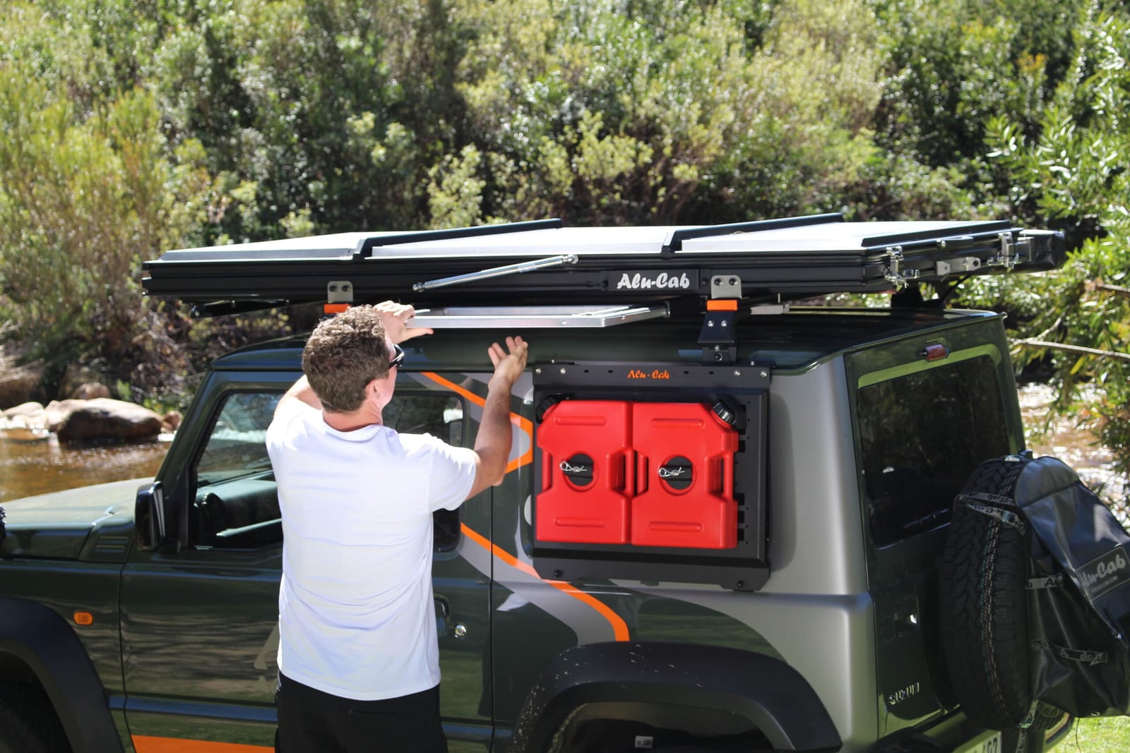 A dark green Suzuki Jimny 3-door beside a river with a closed Alu-Cab LT-50 tent on two load bars and a man sliding out a table under it