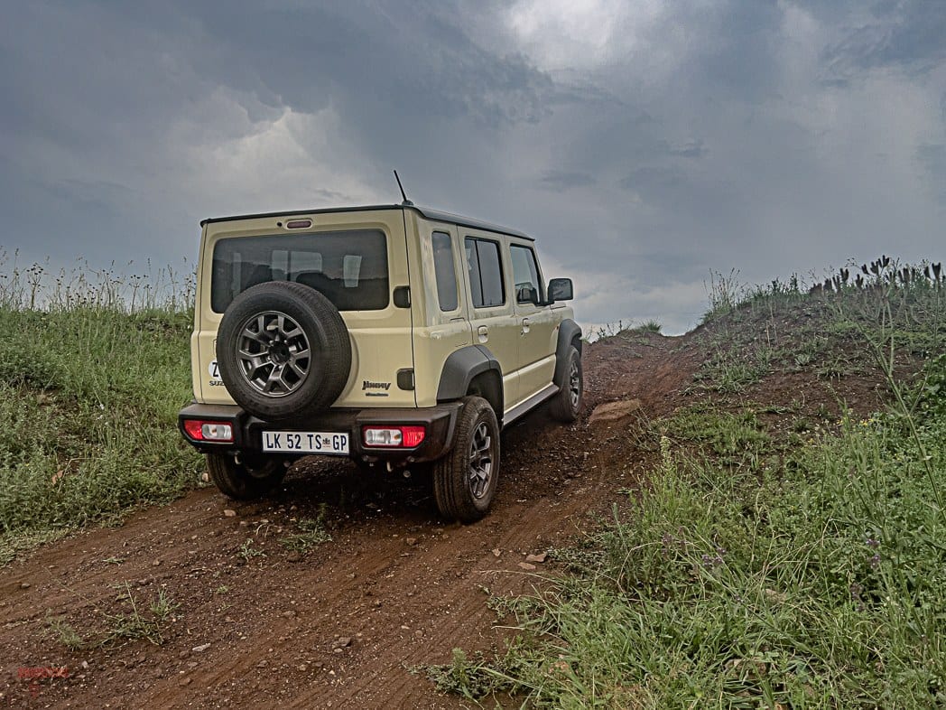 A beige Suzuki Jimny 5-door seen from behind on a red dirt track through long grass under a stormy sky