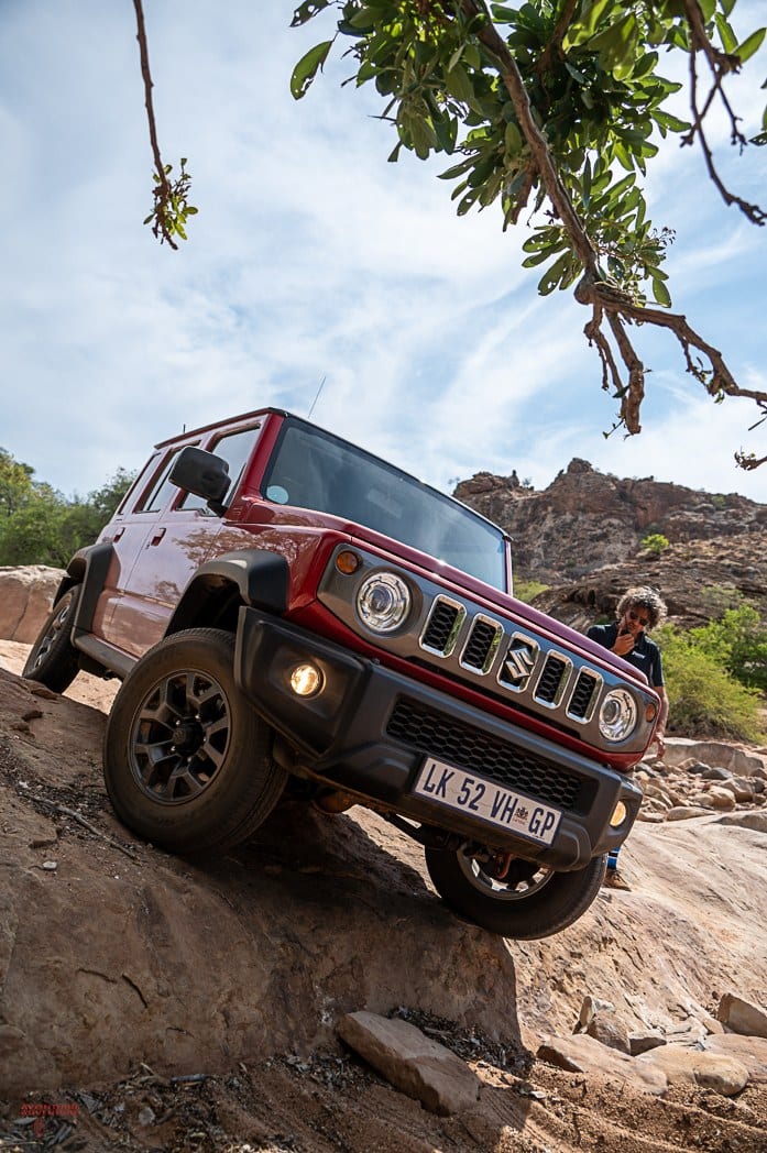 A red Suzuki Jimny 5-door coming nose down off a rock step with one front wheel in the air and a person spotting beside it