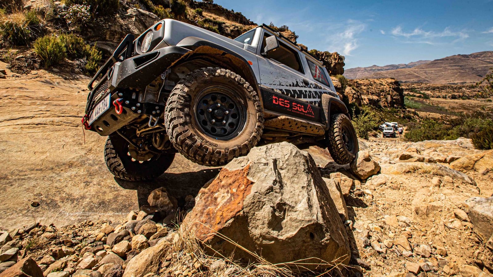 A silver Suzuki Jimny 3-door with a steel front bumper, winch, rock sliders, roof rack and mud-terrain tyres, one front wheel up on a boulder on a rocky mountain track