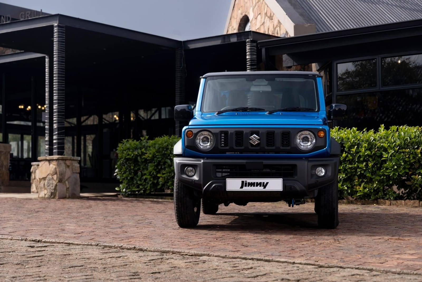 A standard blue Suzuki Jimny 3-door, seen head on, parked on brick paving in front of a stone building