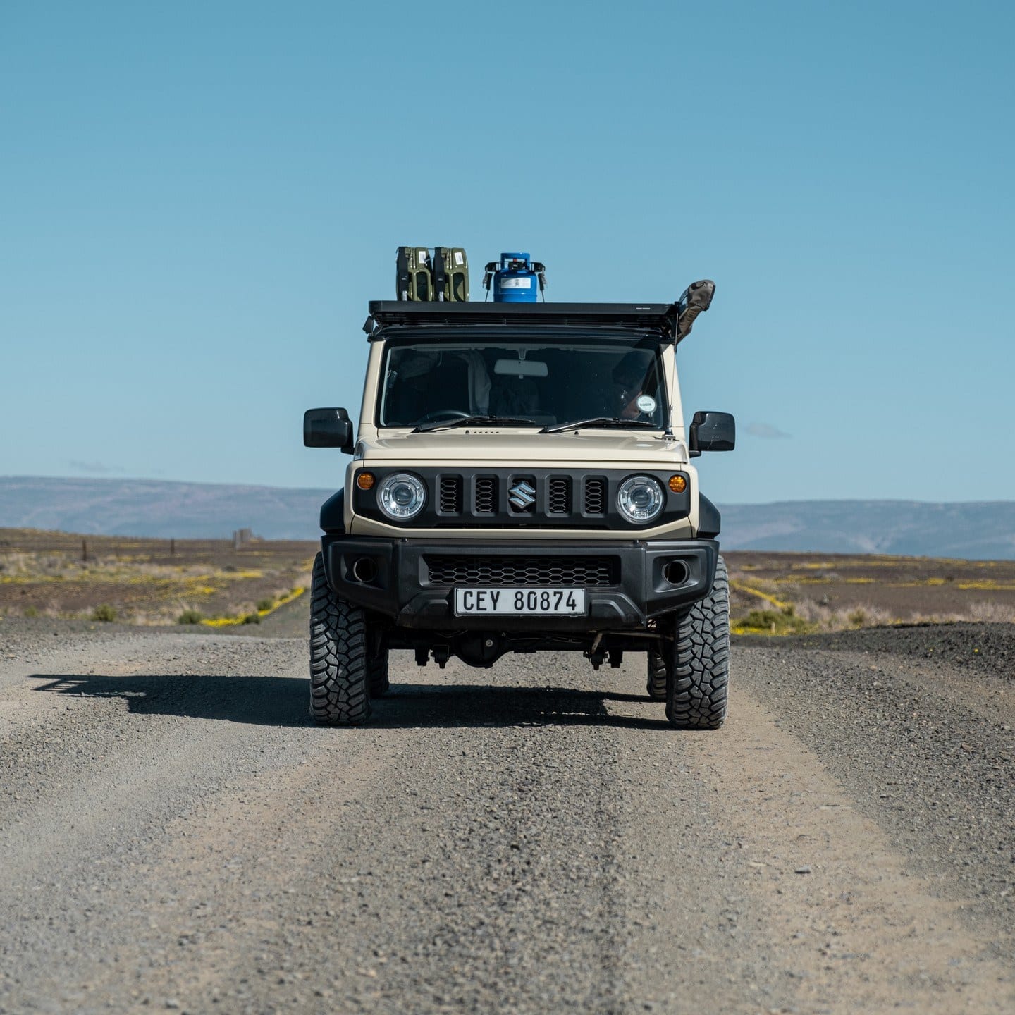 A beige Suzuki Jimny 3-door on a gravel road with a roof rack, jerry cans and all-terrain tyres