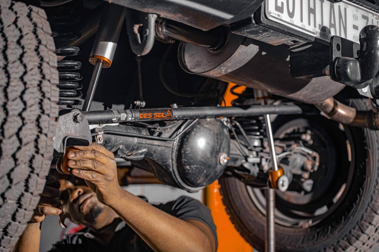 A fitter's hands tightening an orange adjustable bar at the rear axle of a Suzuki Jimny, with the shock absorber and coil spring in view