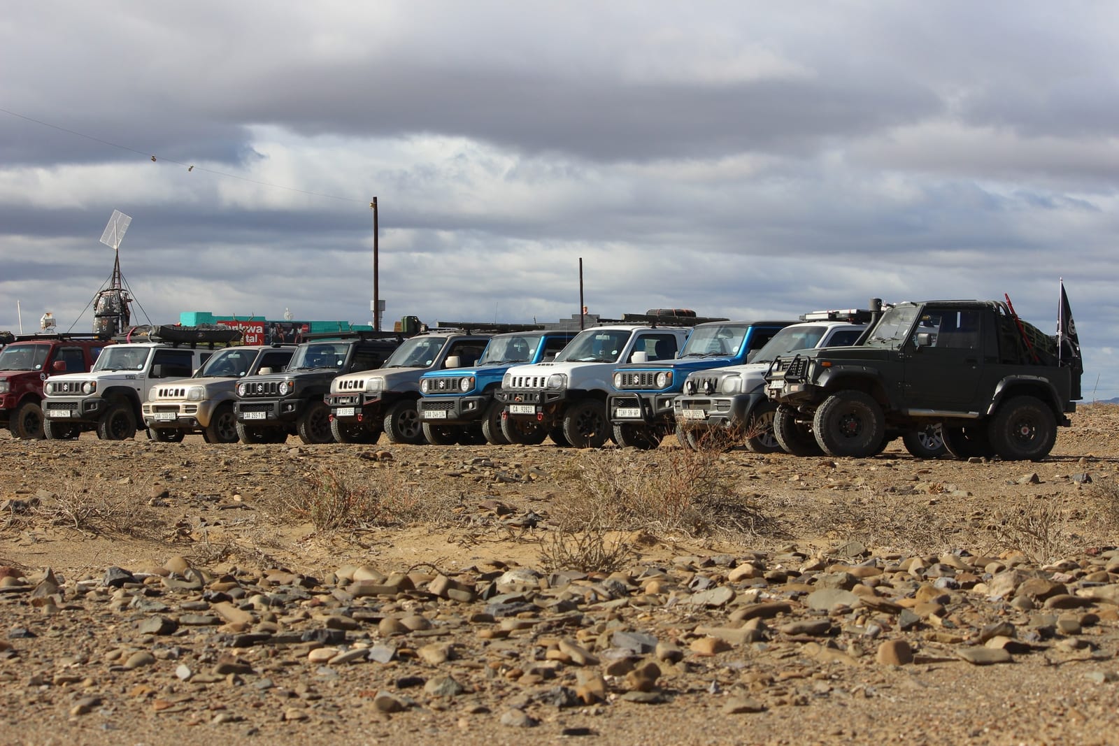 Eleven Suzuki Jimnys parked in a row on stony ground, from nearly stock to heavily built, with an old Suzuki SJ at the end