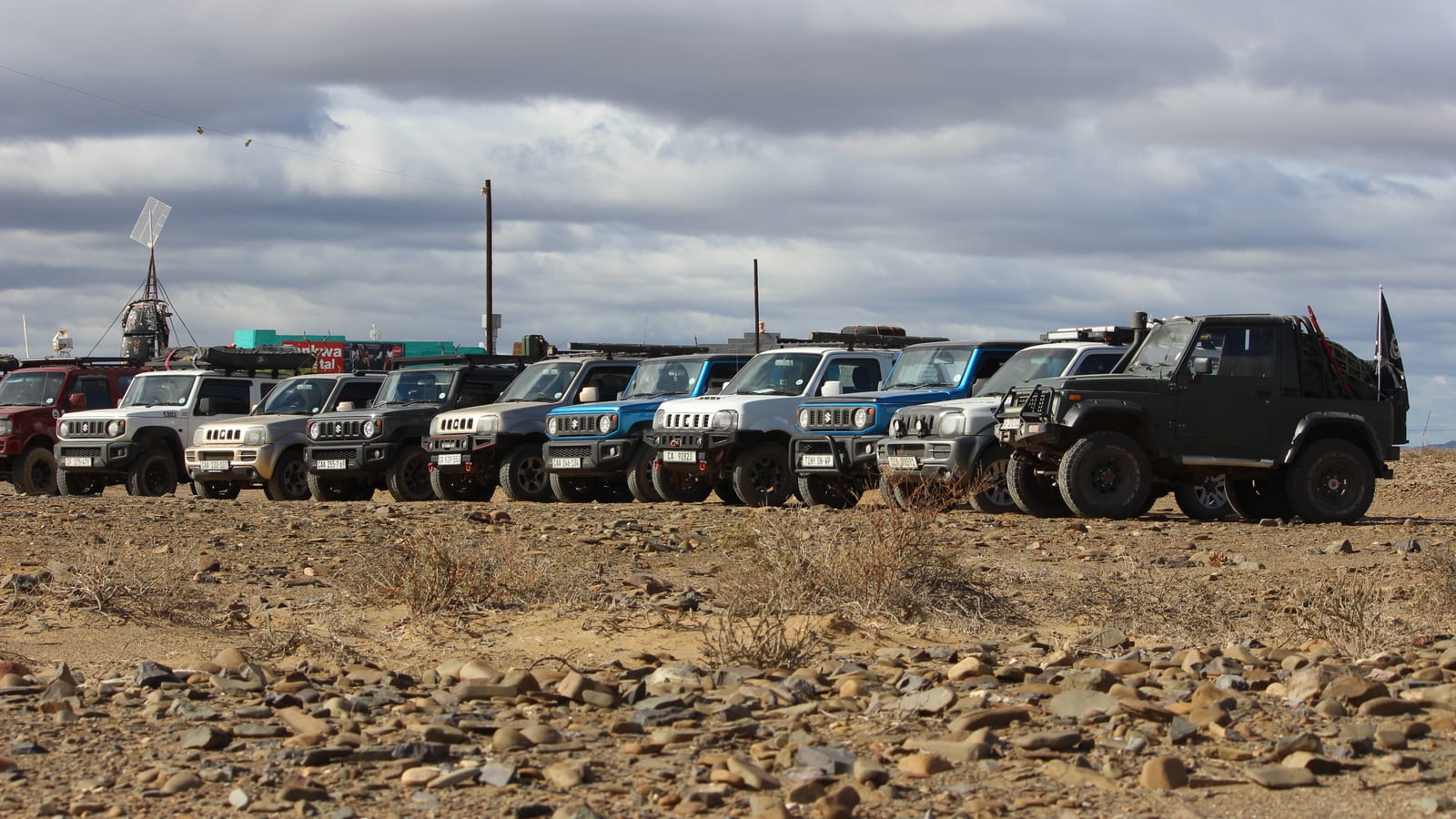 A long line of older and current Suzuki Jimnys parked on stony ground at a Tankwa Karoo meet under a grey sky