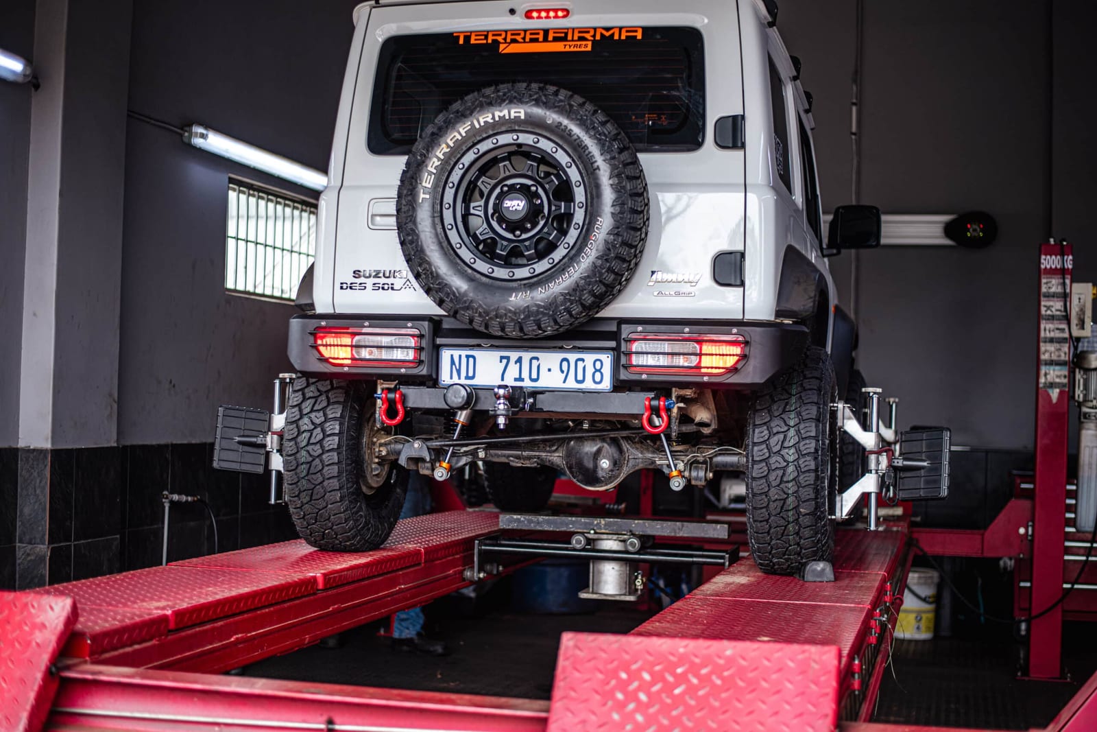 A white current Suzuki Jimny 3-door on a wheel alignment rig in a workshop, seen from behind