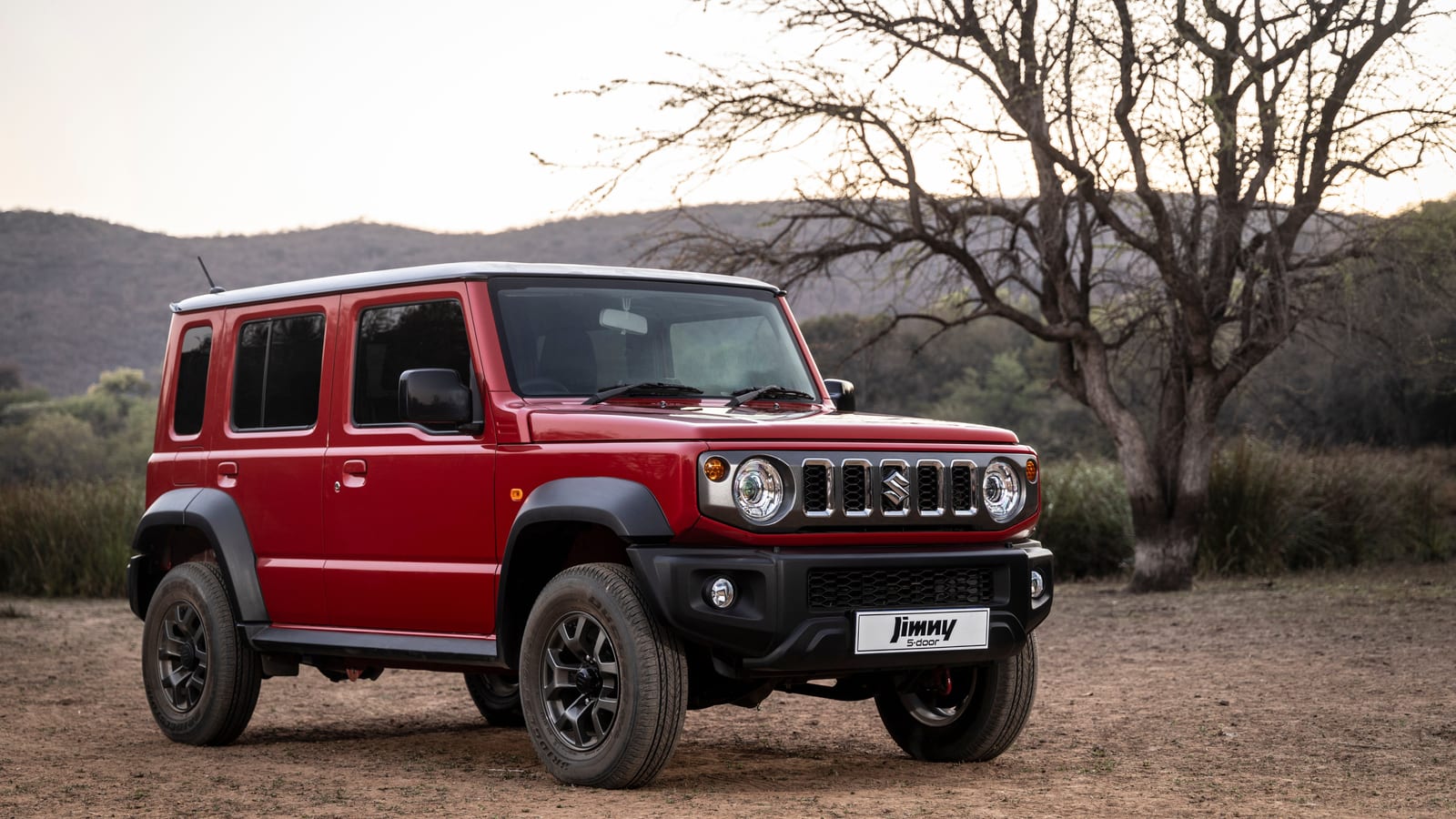 Red Suzuki Jimny 5-door GLX parked on dry bushveld with a bare tree and hills behind