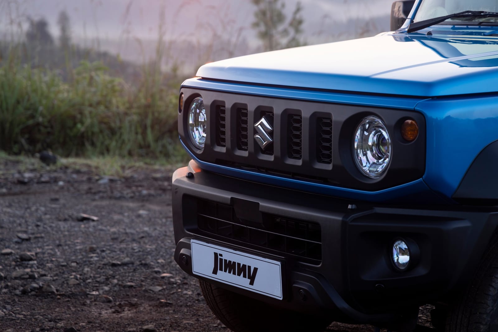 Close-up of the front of a blue Jimny with a round LED projector headlight, the grille and a fog lamp in the bumper