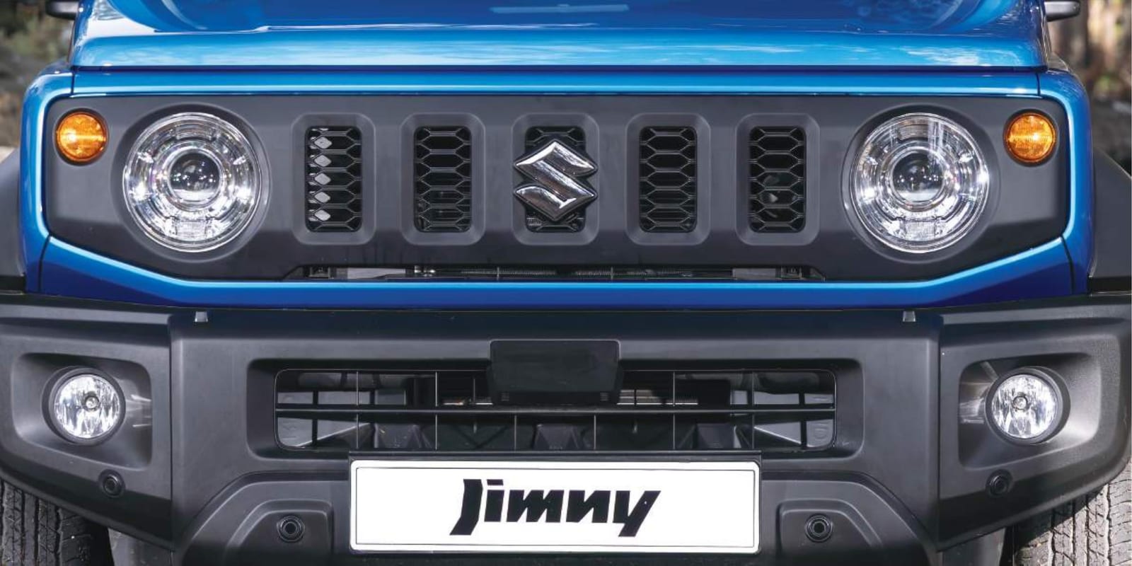 The front of a blue 2026 Jimny 3-door with round LED headlights, fog lamps and four small round sensors in the bumper
