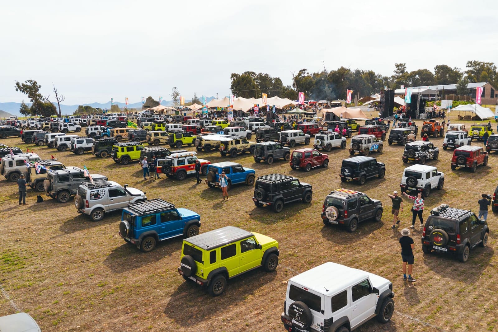 Hundreds of 3-door and 5-door Jimnys in many colours parked on grass at an outdoor event, with tents and a stage behind