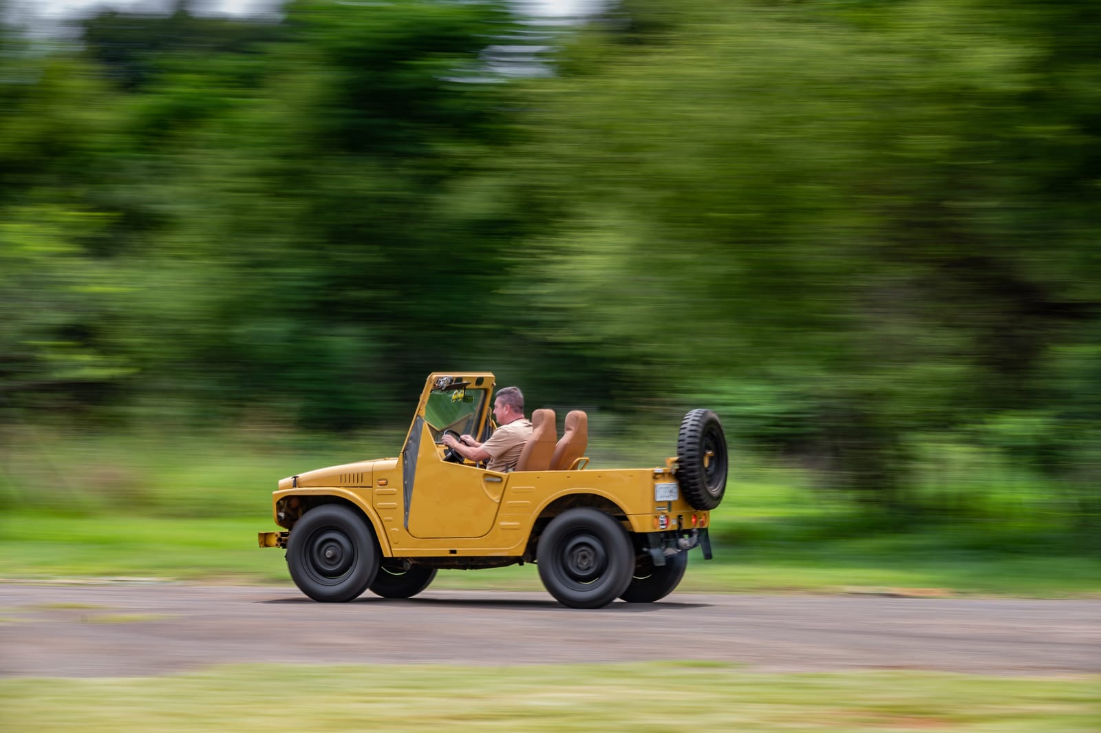 A mustard yellow Suzuki LJ50 soft-top with no roof, driving fast past green trees