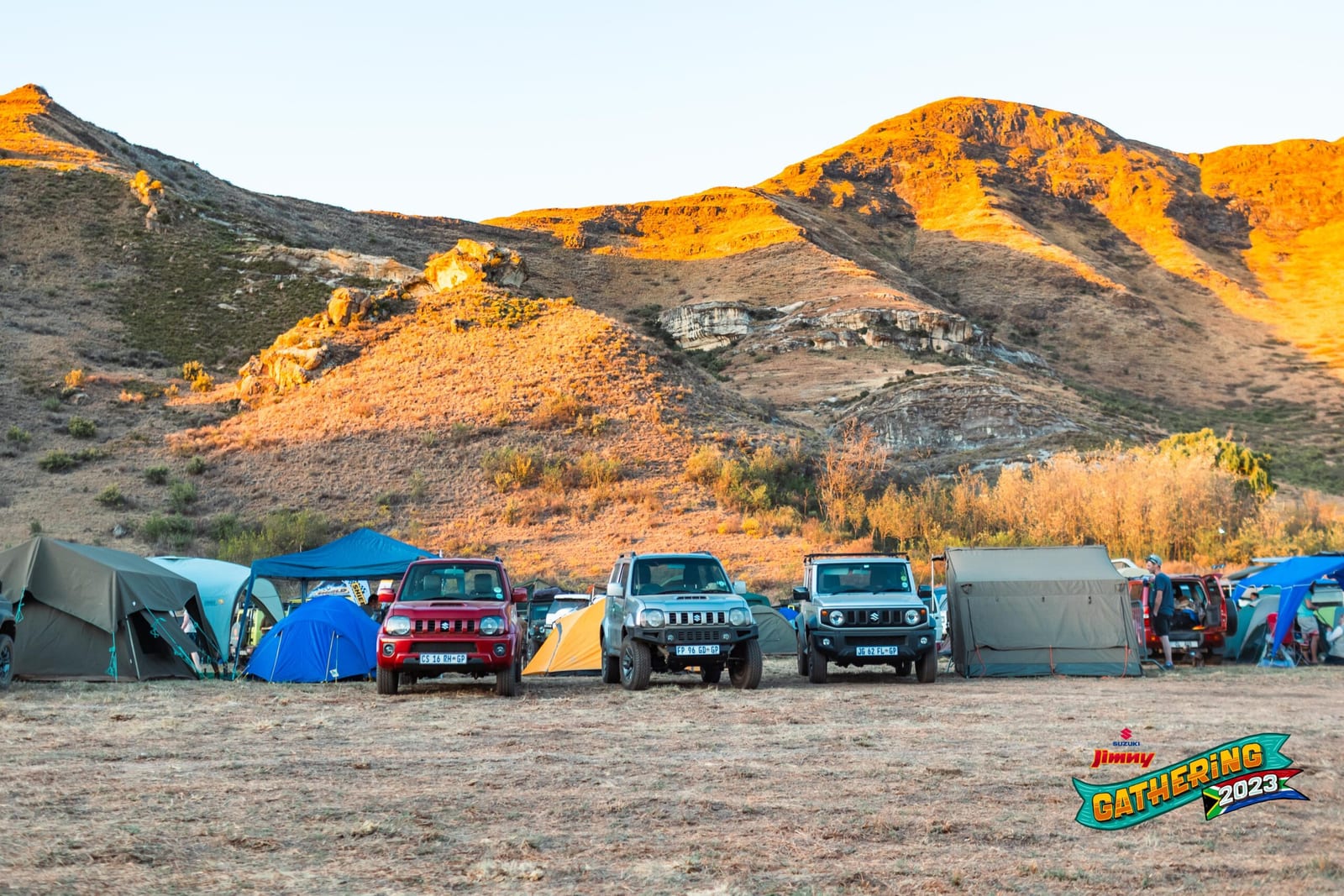 A red and a silver older Suzuki Jimny parked beside a current Jimny among tents, below orange sandstone hills at golden hour