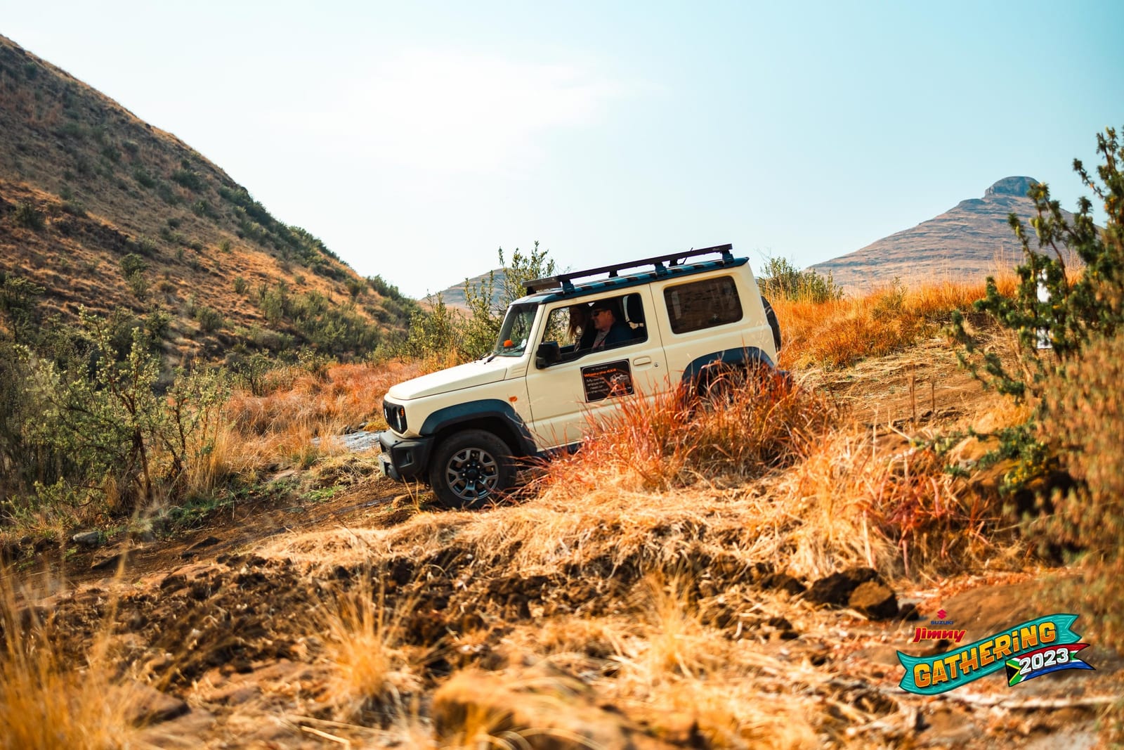 An ivory current Suzuki Jimny 3-door with a roof rack climbing a rutted track through dry grass and bush