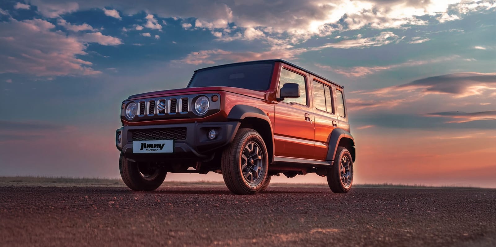 A red Suzuki Jimny 5-door on a flat gravel plain at sunset