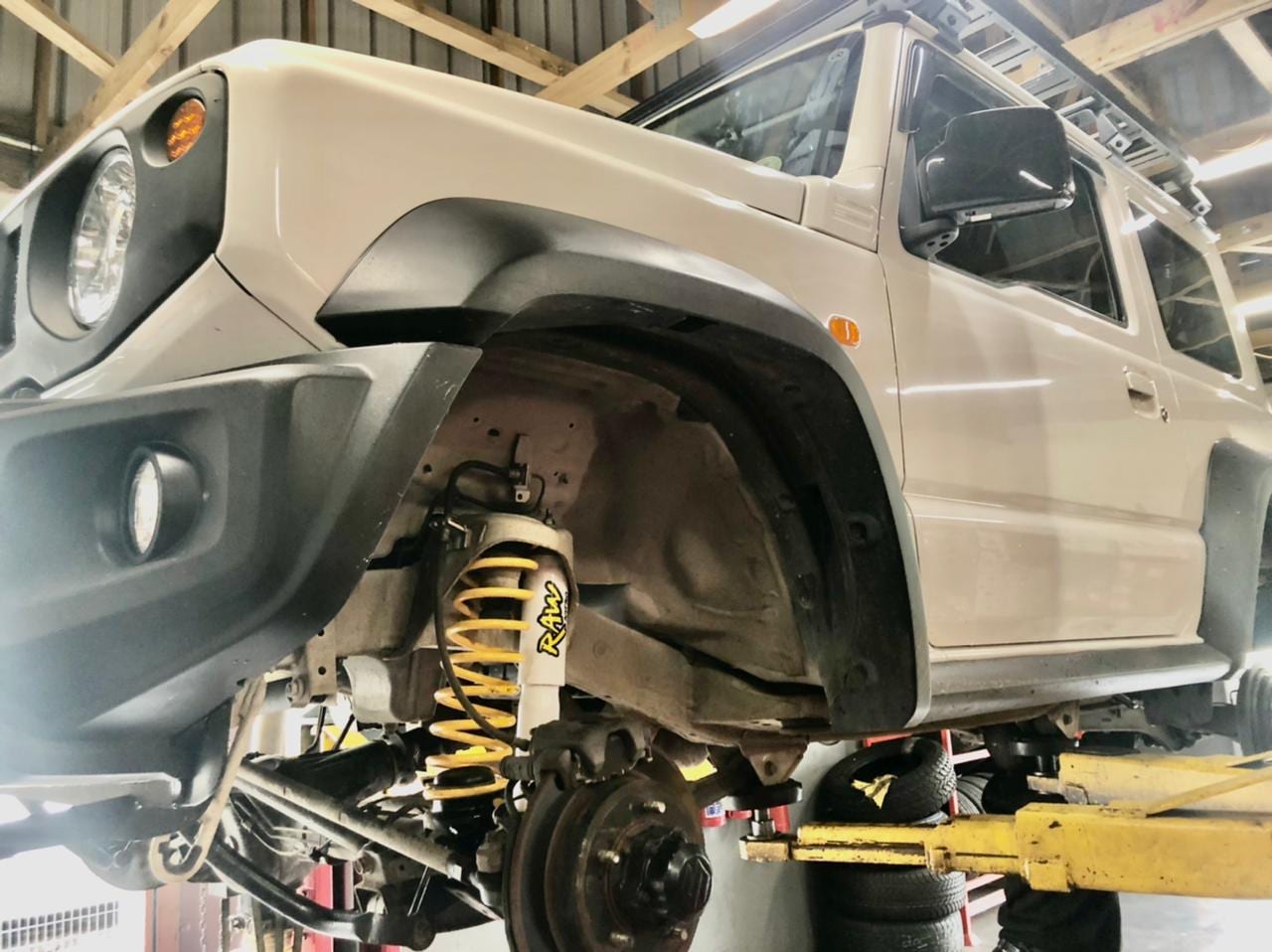 A current Suzuki Jimny up on a workshop hoist with a front wheel off and a yellow coil spring and shock fitted