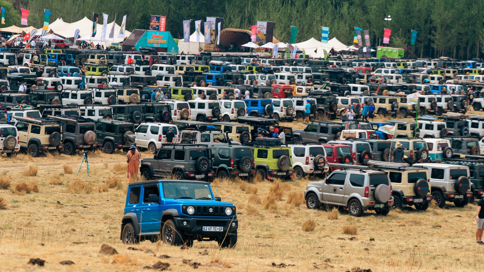 A blue current Suzuki Jimny 3-door and a silver older Jimny parked in front of hundreds of Jimnys of every age at the 2023 Jimny Gathering near Clarens