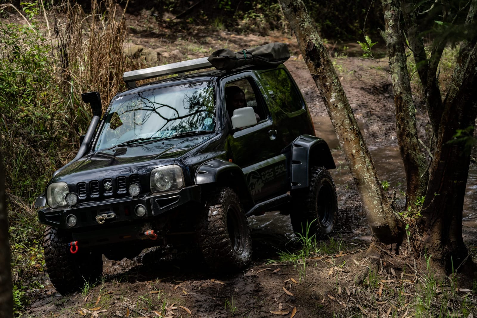 A black older Suzuki Jimny with a snorkel and mud tyres climbing out of a muddy stream between trees