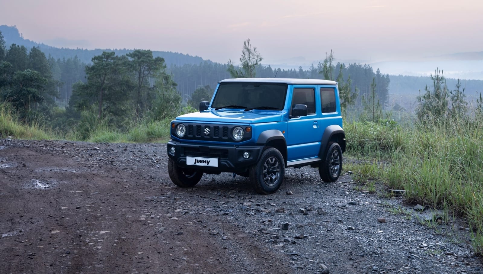 A standard blue Suzuki Jimny 3-door with a black roof on a wet gravel road with pine trees and misty hills behind it