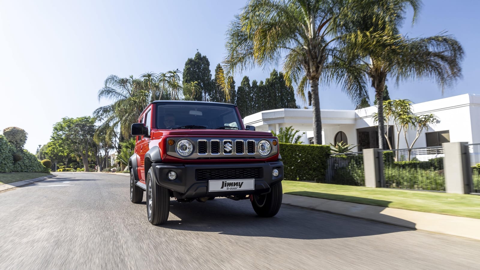 A new red Suzuki Jimny 5-door with a black roof driving down a sunny suburban street lined with palm trees