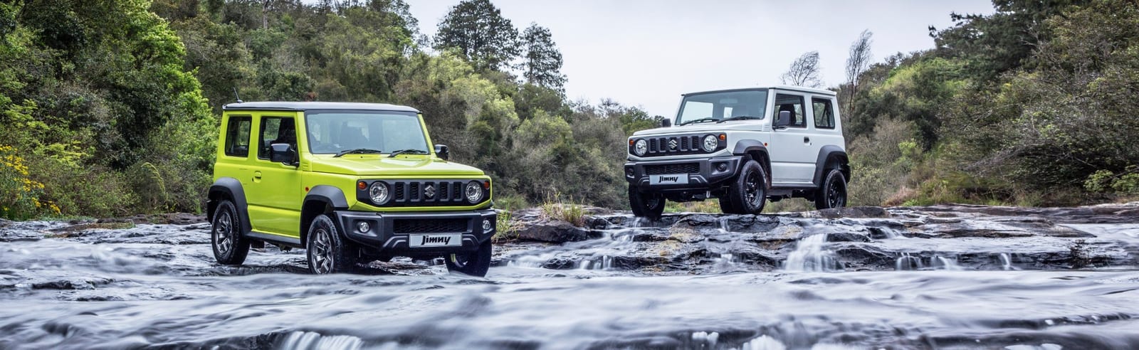 Two standard current Suzuki Jimny 3-doors, one lime green and one white, standing in a shallow rocky river with forest behind