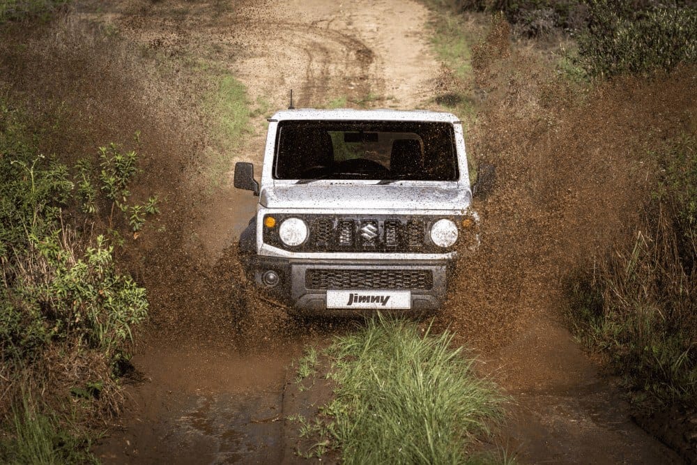 A white standard Suzuki Jimny 3-door driving head-on through mud on a two-track path through the bush