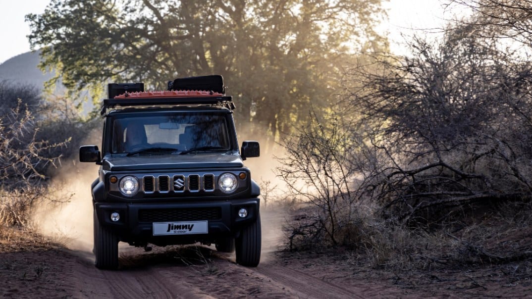 A dark grey Suzuki Jimny with a loaded roof rack driving head-on down a dusty bush track at golden hour