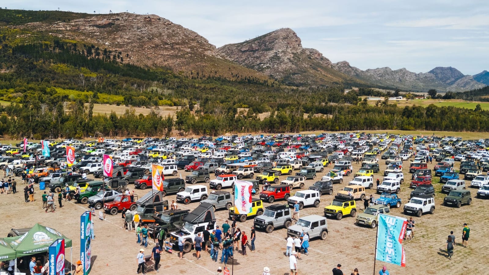 Hundreds of Suzuki Jimnys parked on a field at Jimny Gathering 2025 in the Western Cape, with crowds, flags and mountains behind